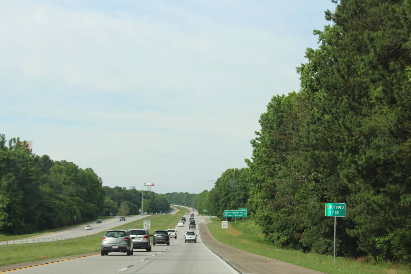 A green 'FLOWERY BRANCH CITY LIMIT' sign stands on the right shoulder of a four-lane divided interstate lined with tall pines and hardwoods; cars drive away toward an exit sign reading 'Spout Springs Rd / Flowery Branch', a 70 mph speed-limit sign stands on the shoulder, and billboards rise above the trees under a pale overcast sky.