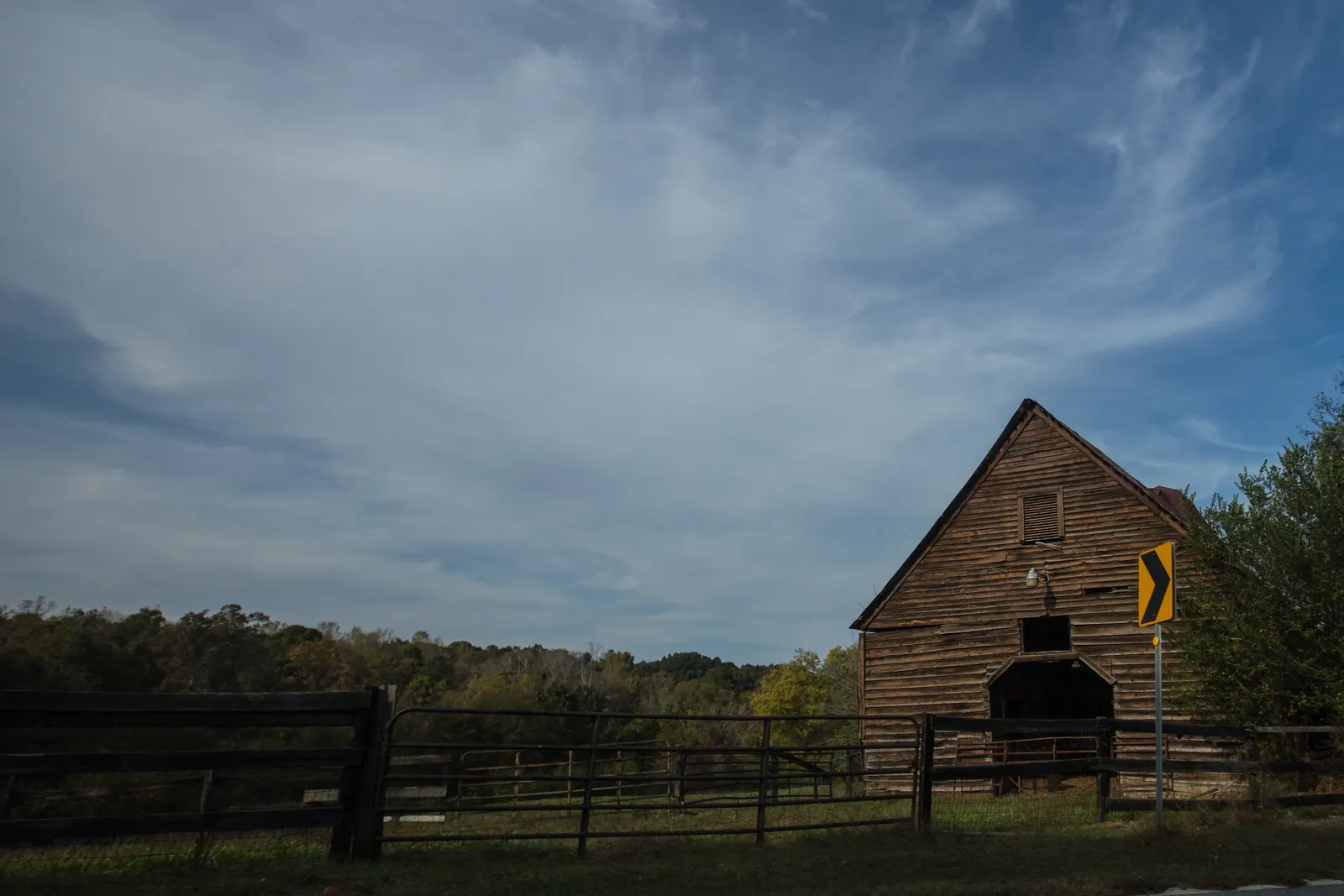 A weathered wooden gabled barn with a dark open doorway and a louvered vent stands at the right edge of a pasture behind black board fencing and metal gates, beside a yellow chevron curve sign; a wooded ridge with early autumn colour runs along the horizon under a wide sky streaked with wispy cirrus cloud.