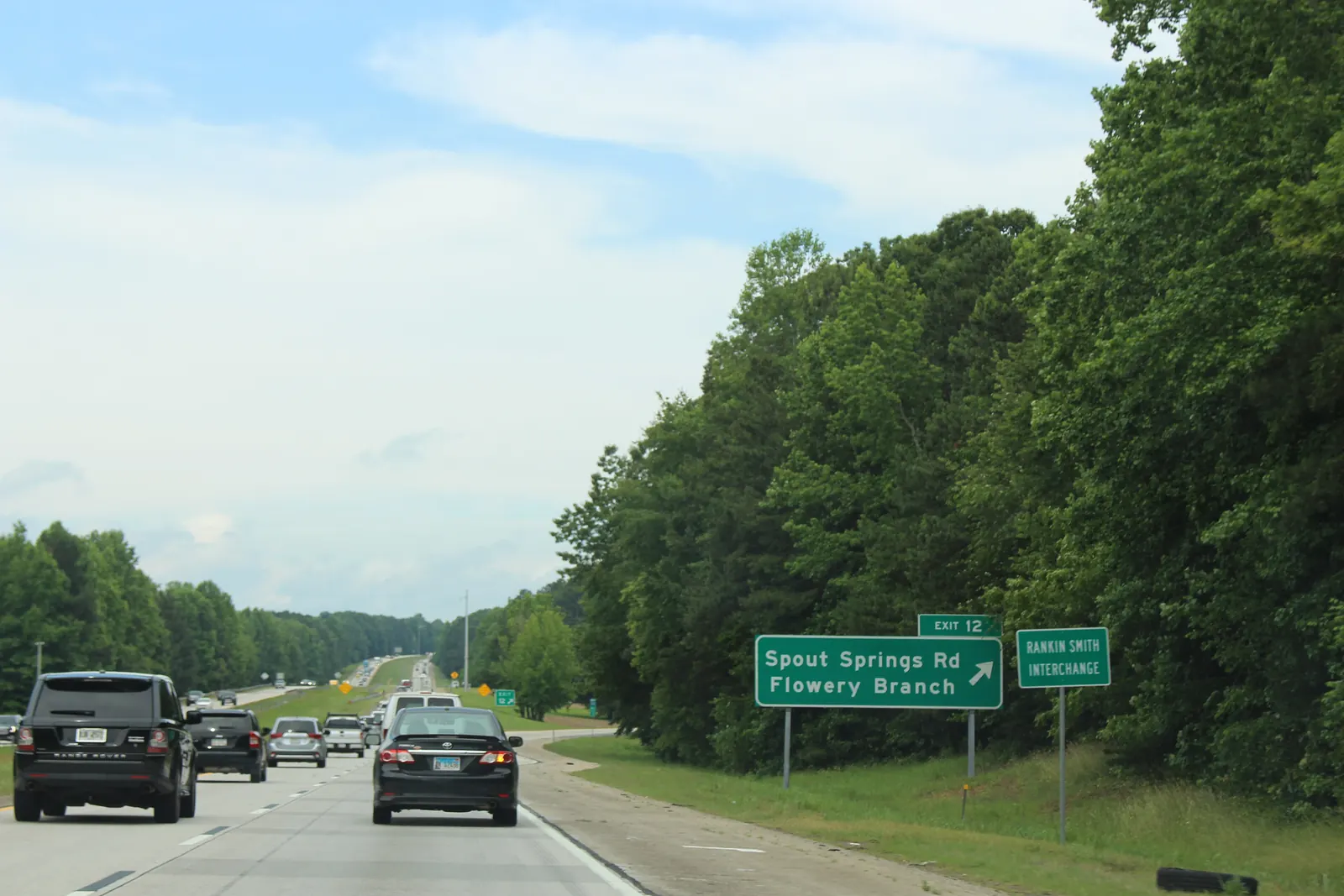Cars, including a black SUV and a black sedan, travel a multi-lane interstate past a green exit sign reading 'EXIT 12 - Spout Springs Rd - Flowery Branch' with an arrow, next to a smaller sign reading 'RANKIN SMITH INTERCHANGE', set against a dense wall of green trees under a partly cloudy sky.