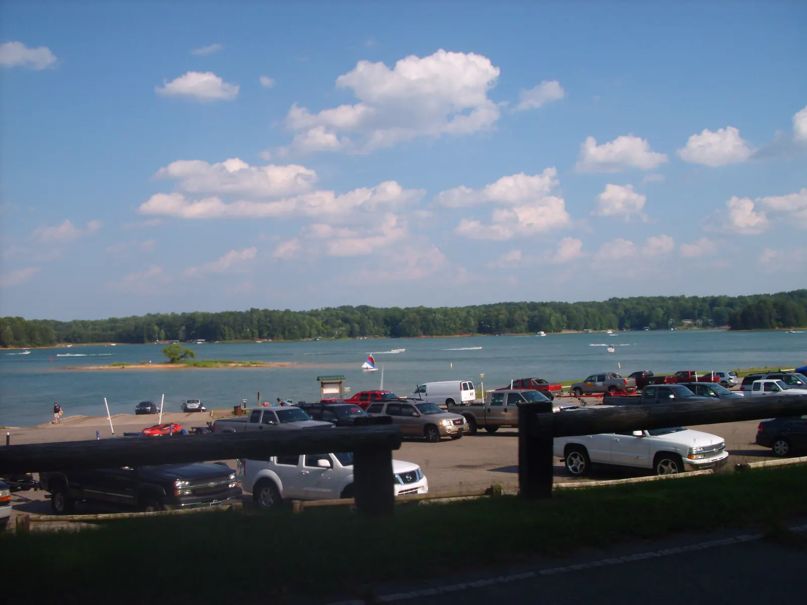 Beyond a wooden rail fence and a parking lot full of pickup trucks with boat trailers, a boat ramp slopes into a broad blue lake dotted with motorboats, a small sailboat and a low grassy islet with one tree, backed by a continuous wooded shoreline under a blue sky with scattered white clouds.