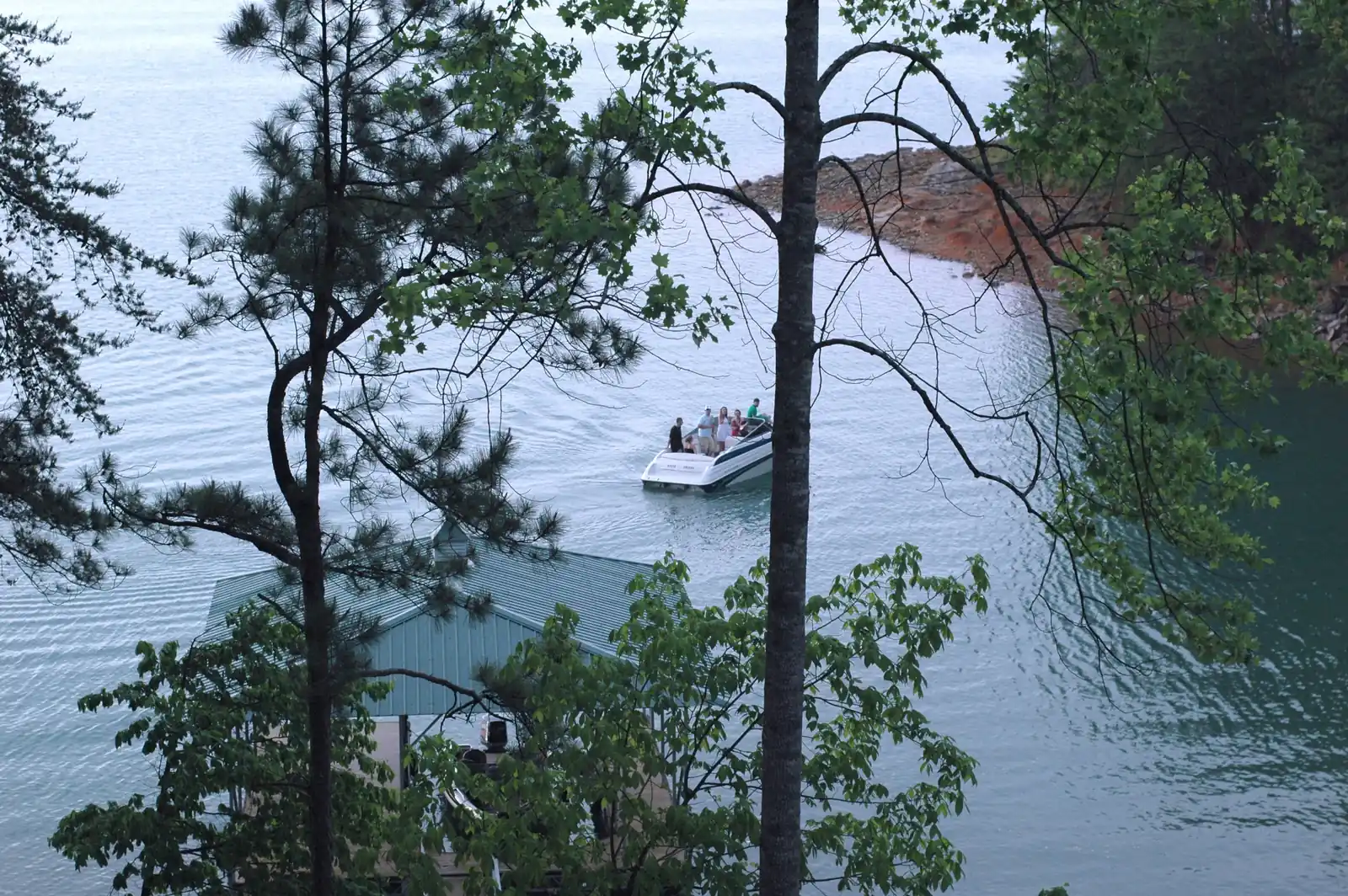 Seen from a hillside through pine and hardwood branches, a white motorboat carrying several people moves across pale blue-green water past a green metal boathouse roof below, toward a point of exposed red clay and rock along the far shoreline.