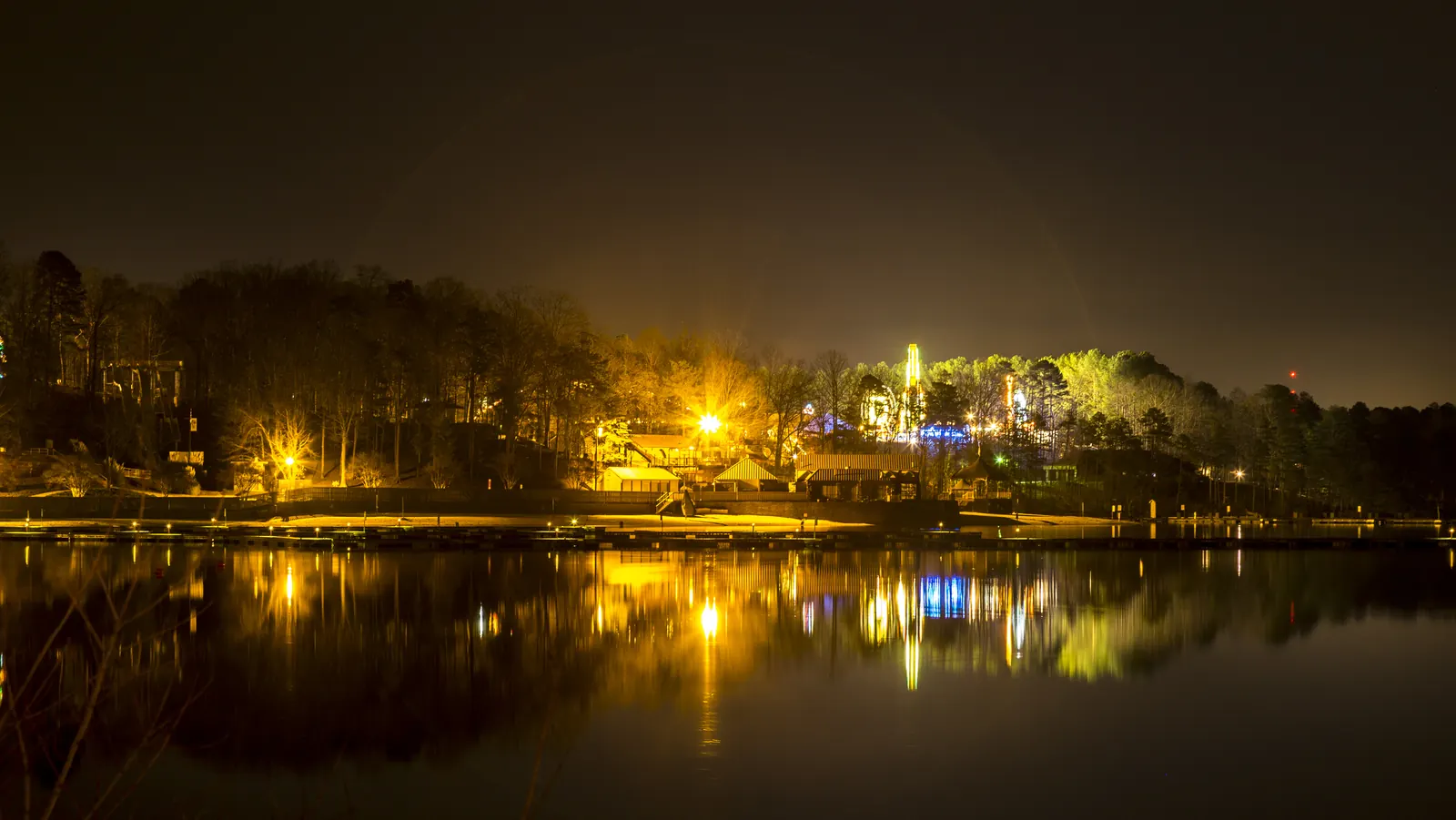 A long night exposure across perfectly still water shows a wooded shoreline lit by warm orange lamps, low buildings, a lit tower and patches of blue and green light, all mirrored in the lake below a dark brown sky.