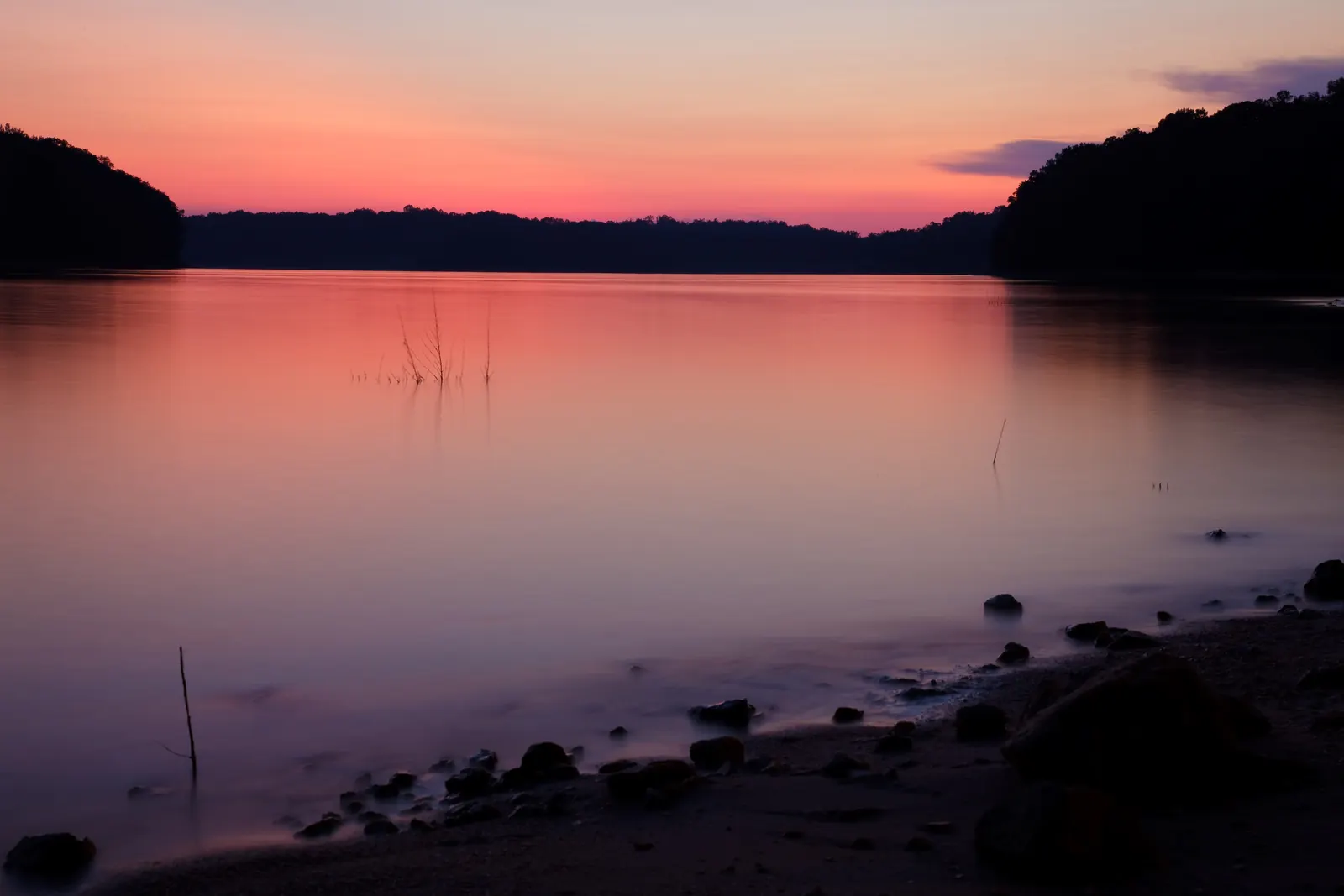 A long exposure at dusk turns glassy lake water pink and violet beneath an orange-to-pink sky; dark wooded hills frame a cove on both sides, a few dead stems poke from the water, and smooth dark stones dot a sandy beach in the foreground.