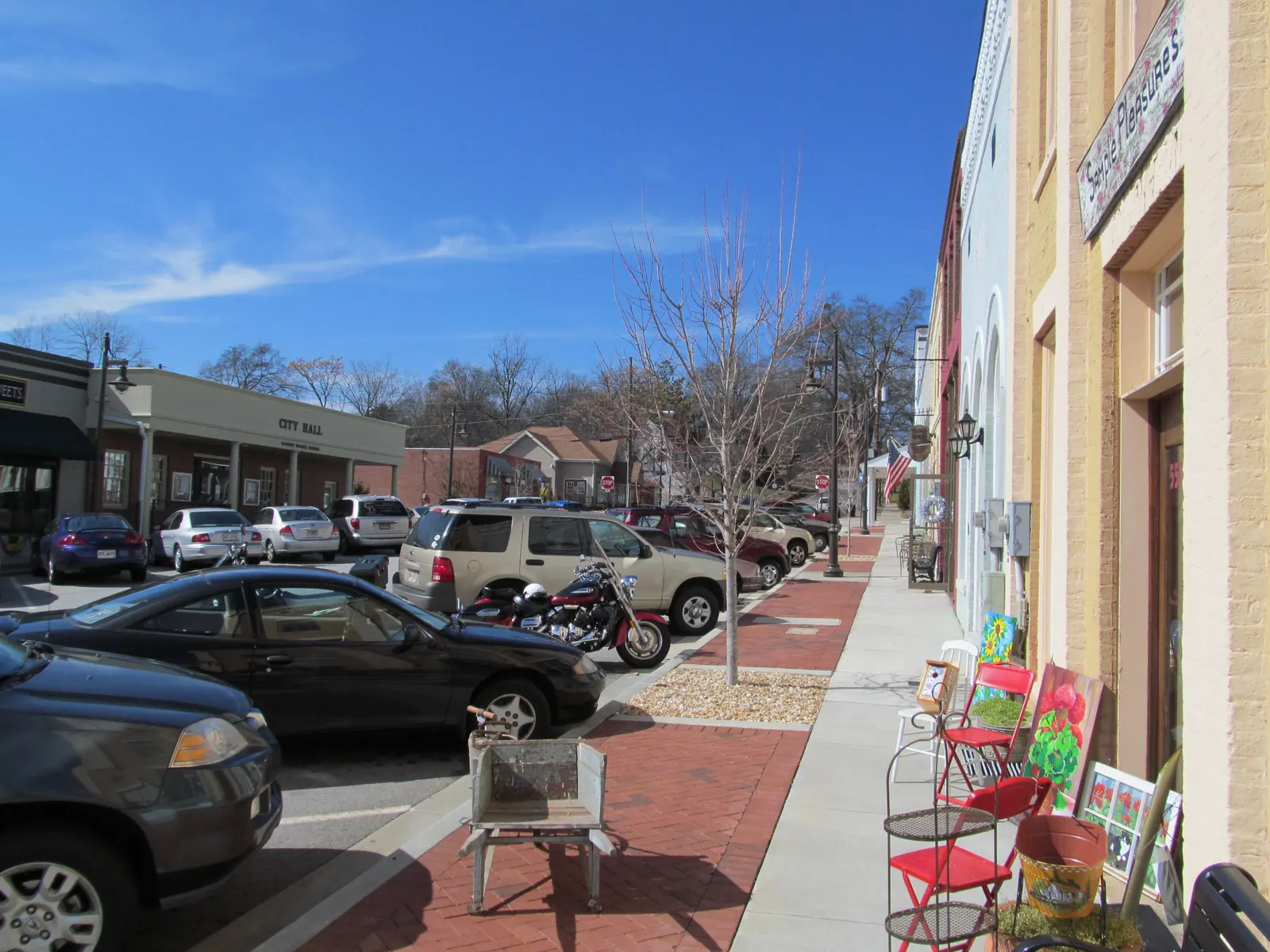 Looking down a sunny brick-paved sidewalk past cream and pale-blue brick storefronts with arched windows, an American flag and a hanging sign reading "Simple Pleasures"; red metal chairs and painted canvases sit outside a shop on the right, cars and a motorcycle are angle-parked along the street, and across the street a low brick building carries a "CITY HALL" sign under a deep blue sky with bare winter trees.